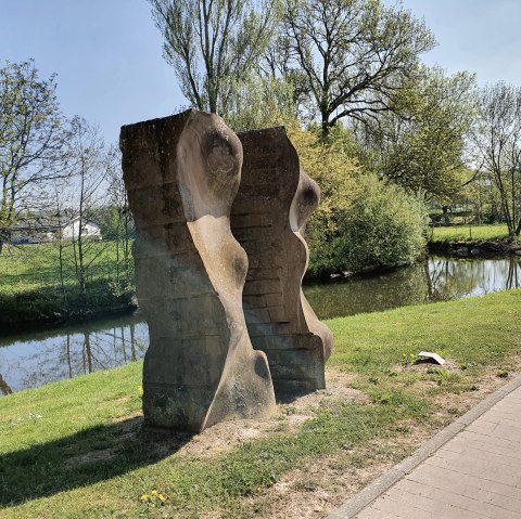 Steinskulptur am Flussufer, umgeben von grüner Landschaft und Bäumen. Ein klarer Himmel und ein Weg sind ebenfalls sichtbar., © TI Bitburger Land, Steffi Wagner