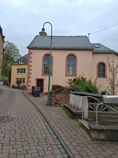 Eine malerische Straße mit einer rosa gefärbten Kirche und einem angrenzenden Gebäude. Die Umgebung ist von Bäumen und einer ruhigen Atmosphäre geprägt.