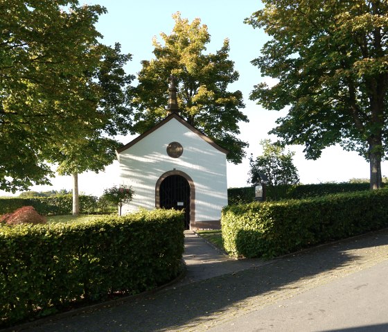 Kleine weiße Kapelle mit braunem Dach, umgeben von grünen Bäumen und Hecken, bei sonnigem Wetter., © Berscheid