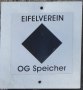 Wanderwegmarkierung des Eifelvereins mit schwarzem Rhombus und Schriftzug 'OG Speicher' auf weißem Hintergrund.