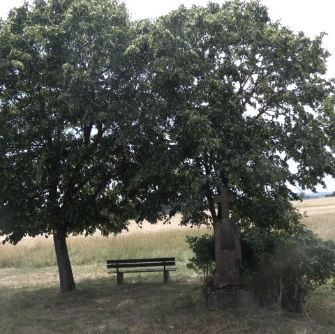 Two large trees provide shade over a bench and a stone cross on a country lane. Fields and a cloudy sky can be seen in the background., © TI Bitburger Land