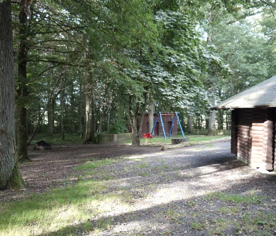 Une aire de jeux dans la forêt avec une balançoire et une cabane à barbecue. Les alentours sont entourés d'arbres et il y a un espace ouvert avec de l'herbe., © Berscheid