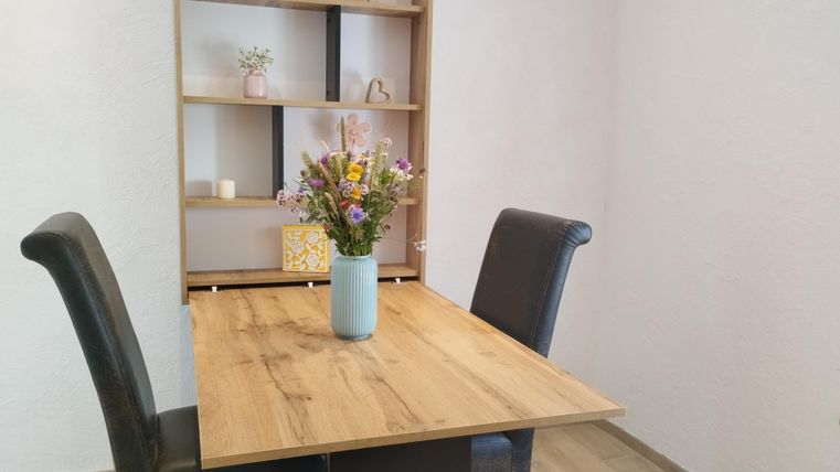 Ein gemütlicher Essbereich mit einem Holztisch und zwei schwarzen Stühlen. Auf dem Tisch steht eine Vase mit bunten Blumen.