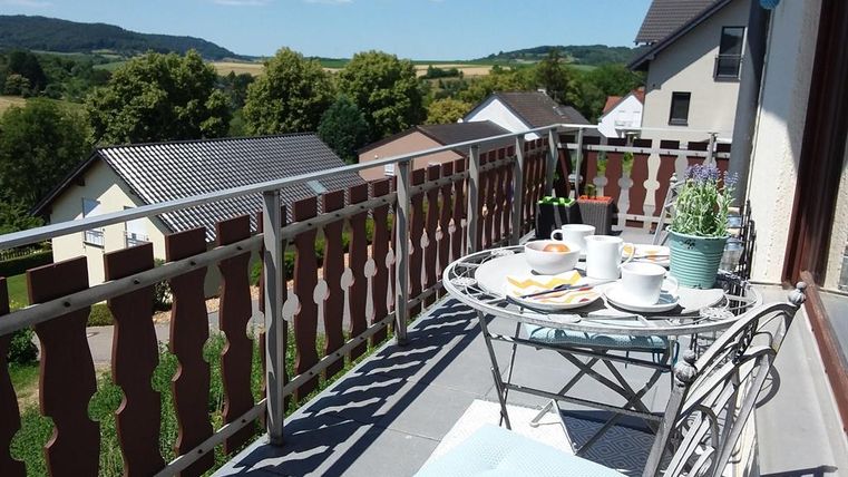 Un balcon accueillant avec une table, garnie d'ustensiles de petit déjeuner et de tasses colorées. À l'arrière-plan, un paysage verdoyant s'étend sous un ciel bleu clair.