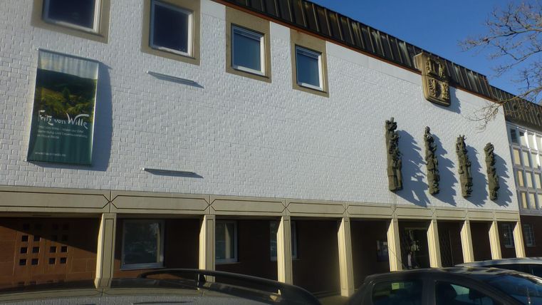 Ein modernes Gebäude mit einer hellen Fassade und mehreren Fenstern. Im Vordergrund stehen geparkte Fahrzeuge.