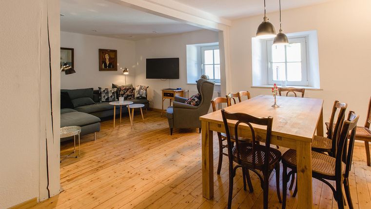 Ein helles und einladendes Wohnzimmer mit einem großen Esstisch und bequemen Sofas. Der Boden ist aus Holz, und die Wände sind schlicht und neutral.