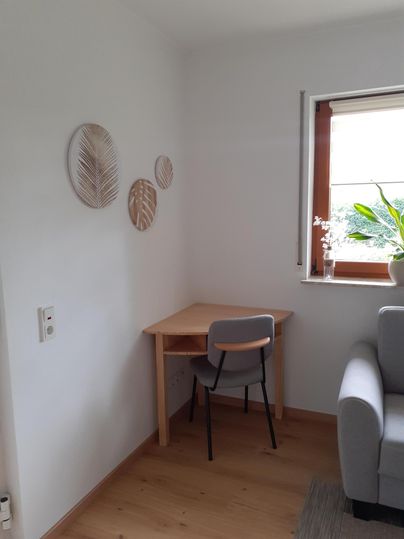 Een lichte kamer met een kleine tafel en een stoel in de hoek. Aan de muur hangen decoratieve bladeren, en een raam met planten zorgt voor natuurlijke verlichting.