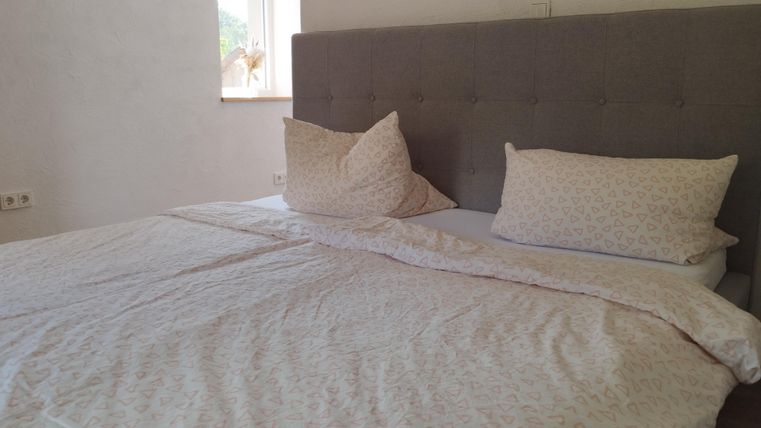 Ein ordentliches Bett mit grauem Kopfteil und blauen Kissen. Die Bettwäsche hat ein dezentes Muster und die Fenster lassen natürliches Licht herein.