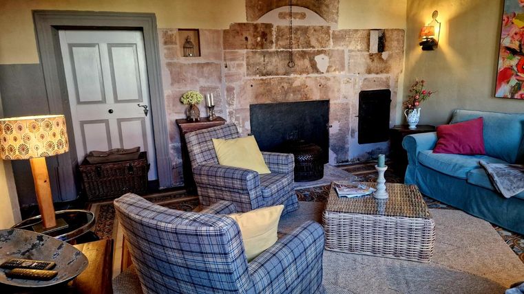 Un salon confortable avec une cheminée, deux fauteuils et un canapé. Les murs sont peints dans des couleurs chaudes et il y a des éléments décoratifs comme des fleurs et un tapis.