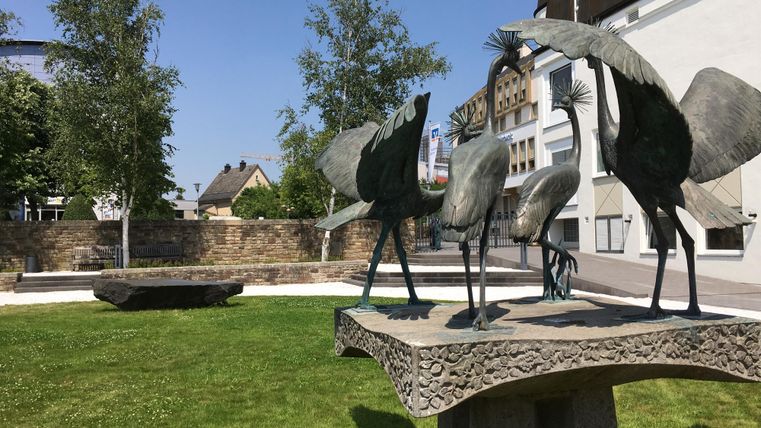 Eine moderne Skulptur von Vögeln steht auf einem Sockel in einer grünen Parklandschaft. Im Hintergrund sind Bäume und Gebäude sichtbar.