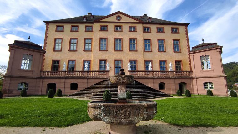 An impressive castle with a beautiful façade and large windows. In the foreground, there is a fountain surrounded by well-maintained grass.