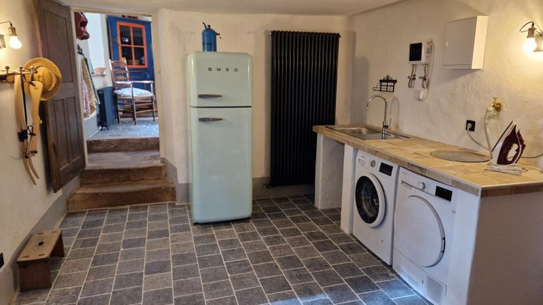 Une cuisine moderne avec des carreaux gris, un réfrigérateur et une machine à laver. À l'arrière-plan, des meubles et une porte menant à une autre pièce sont visibles.