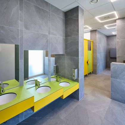 Moderne Badezimmer mit grauen Fliesen und einer auffälligen gelben Waschtischfläche. Die Umgebung ist hell und sauber, mit Trennwänden in verschiedenen Farben.