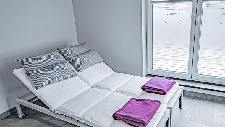 Two white loungers with cushions and purple towels in a bright room. Large window with a view outside.
