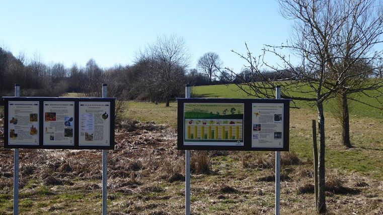 Informationstafeln in einer ländlichen Landschaft mit Bäumen und Wiesen.