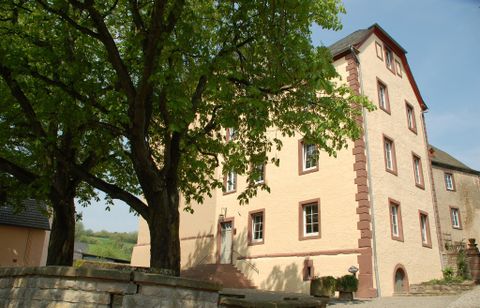 Ein historisches Gebäude mit einem roten und beigen Anstrich, umgeben von Bäumen. Der Himmel ist klar und sonnig.