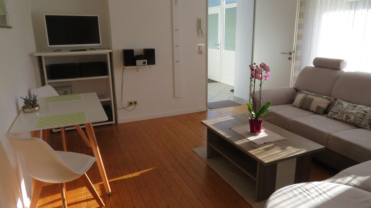 Ein heller Wohnraum mit einem bequemen Sofa und einem Couchtisch. Ein Schreibtisch und ein Fernseher sind ebenfalls sichtbar, während frische Blumen auf dem Tisch stehen.