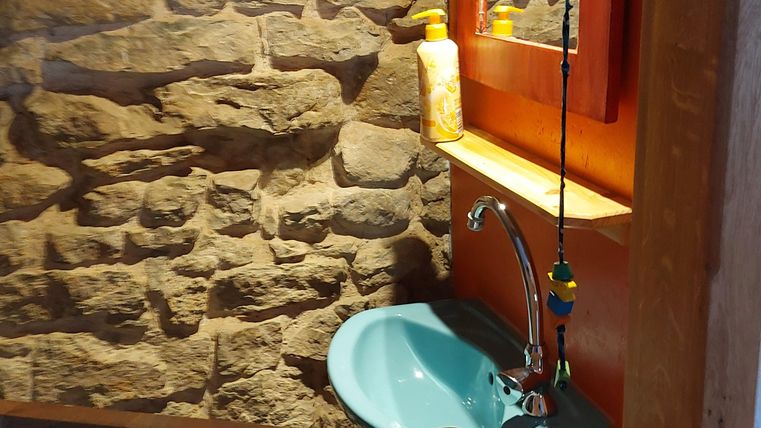 Een kleine wasruimte met een blauwe wastafel en een spiegel. De muren zijn van steen en er is warm licht dat een gezellige sfeer creëert.