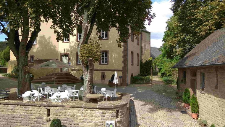 Ein ruhiger Innenhof mit einem historischen Gebäude und Bäumen. Tische und Stühle stehen zum Entspannen im Freien bereit.