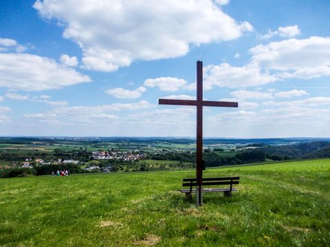 Ein großes Holzkreuz steht auf einer Wiese mit einer Bank davor. Im Hintergrund ist eine weite Landschaft mit einem Dorf zu sehen.