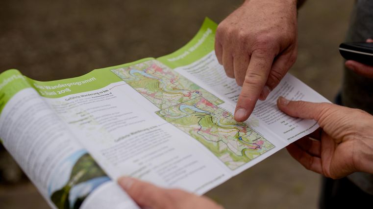 Eine Person zeigt mit dem Finger auf eine Landkarte. Zwei Hände halten die Karte, während die andere Hand ein Smartphone hält.