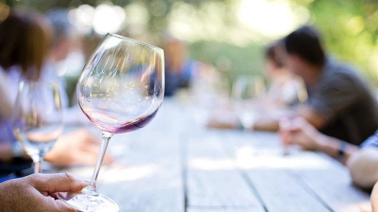 Eine Hand hält ein Weinglas mit rotem Wein. Im Hintergrund sieht man eine Gruppe von Menschen an einem langen Tisch im Freien.
