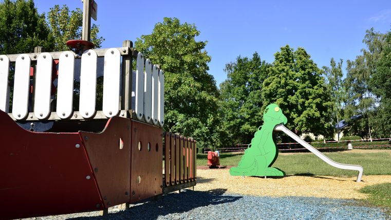 Un terrain de jeu avec un bateau de jeu rouge et un toboggan vert en forme d'animal. En arrière-plan, on peut voir des arbres et un ciel bleu clair.