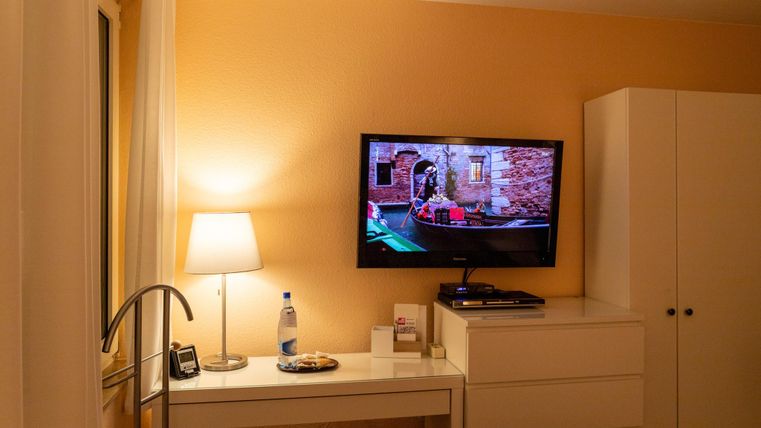 Ein modernes Zimmer mit einem Fernseher an der Wand und einer weißen Kommode. Auf dem Tisch stehen eine Lampe, eine Flasche Wasser und ein paar Snacks.