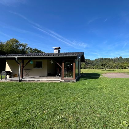 Ein einfaches Haus auf einer Wiese mit grünem Gras. Im Hintergrund sind Bäume und ein klarer, blauer Himmel zu sehen.