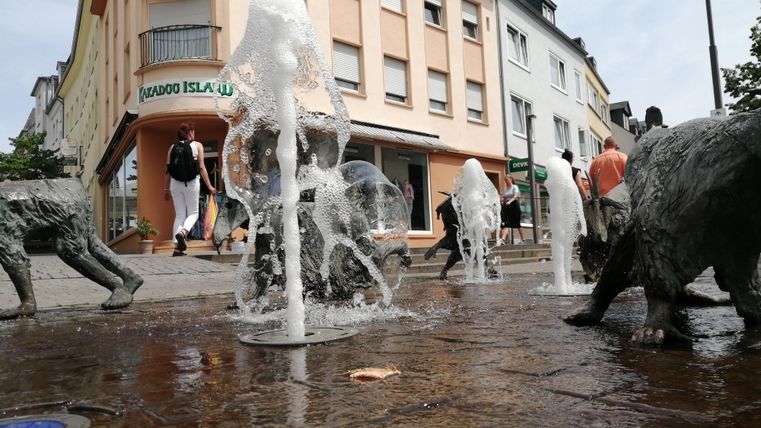 Ein Brunnensystem mit mehreren Wasserfontänen und Tierfiguren ist zu sehen. Im Hintergrund stehen verschiedene Häuser und Passanten gehen vorbei.