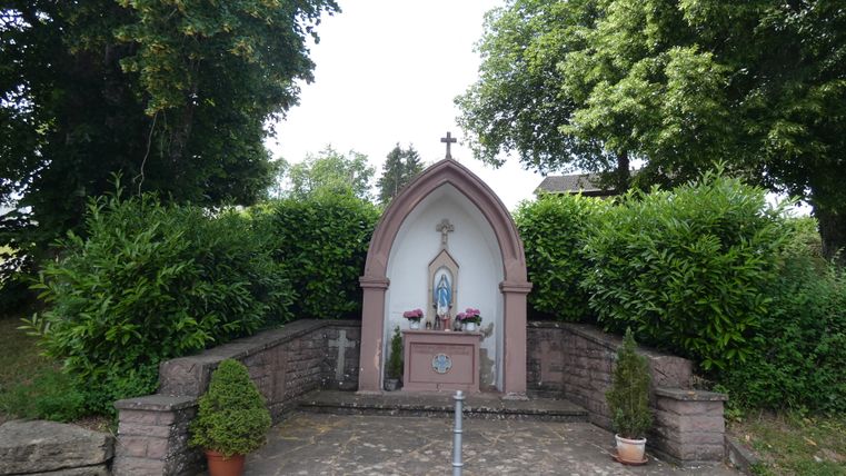 Kleine Kapelle mit Marienstatue, umgeben von Bäumen und Büschen.