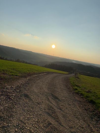 Un chemin traverse un paysage verdoyant, avec un coucher de soleil en arrière-plan. Le ciel est dégagé et l'ambiance est paisible.