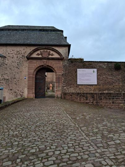 Ein historischer Eingang mit einem großen Tor und einer Steinmauer. Der Platz ist mit Kopfsteinpflaster bedeckt und es gibt eine beschriftete Tafel an der Wand.