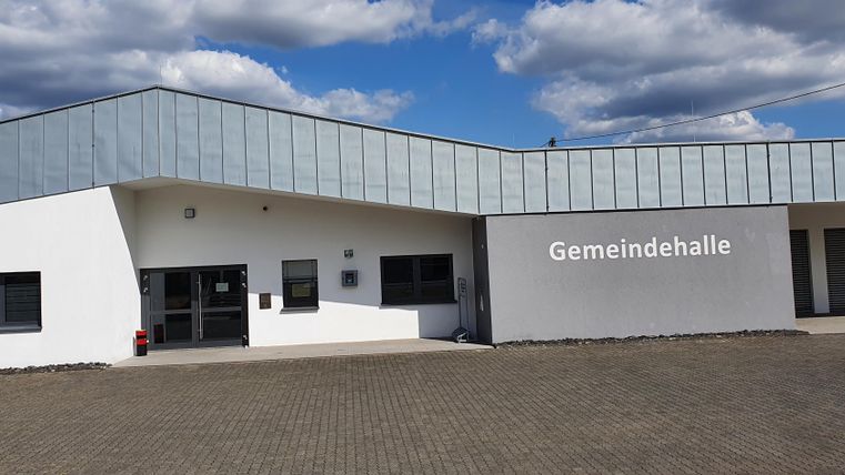 Eine moderne Gemeindehalle mit einer schlichten weißen Fassade und einem hellen Dach. Der Himmel ist blau mit einigen Wolken.