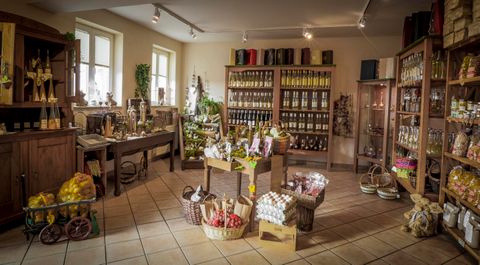 Ein gemütlicher Laden mit Regalen voller Vorräte und Genussmitteln. In der Mitte stehen Körbe und Tische mit verschiedenen Produkten.