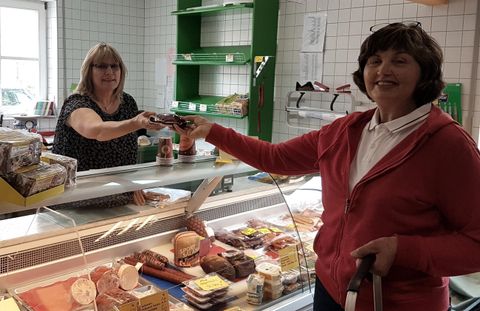 Eine Frau kauft an einer Theke Lebensmittel. Im Hintergrund steht eine Verkäuferin und reicht ihr eine Ware.