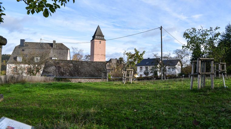 Ein ruhiger Platz mit einer Wiese und historischen Gebäuden im Hintergrund. Im Zentrum steht ein Turm, umgeben von Bäumen und einer klaren blauen Himmel.
