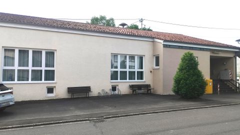 A simple building with a white facade and several windows. Benches are outside and a small tree is visible.