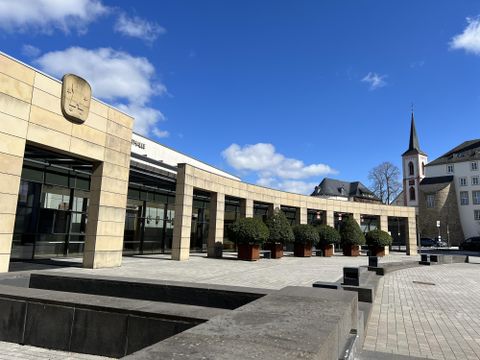 Un bâtiment moderne avec une façade en verre et des plantes devant l'entrée. En arrière-plan, on voit un clocher et un ciel bleu clair.