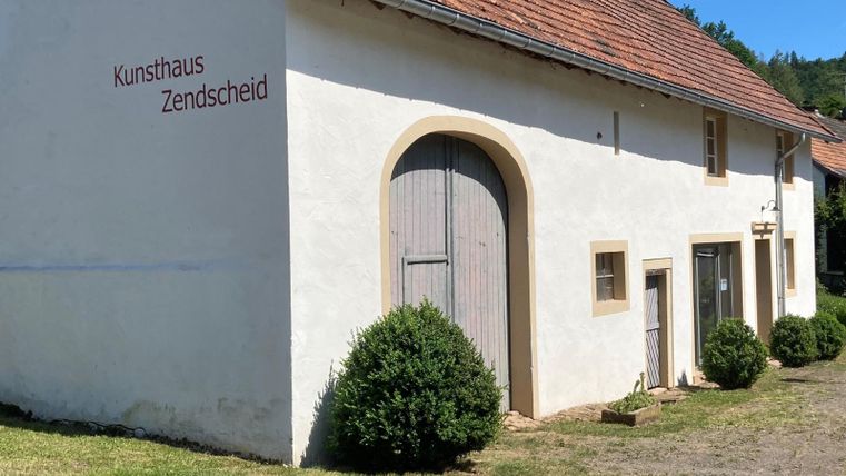 Ein Kunsthaus mit einer weißen Fassade und einem roten Dach. Der Eingang ist mit einer großen Holztür und kleinen Sträuchern dekoriert.