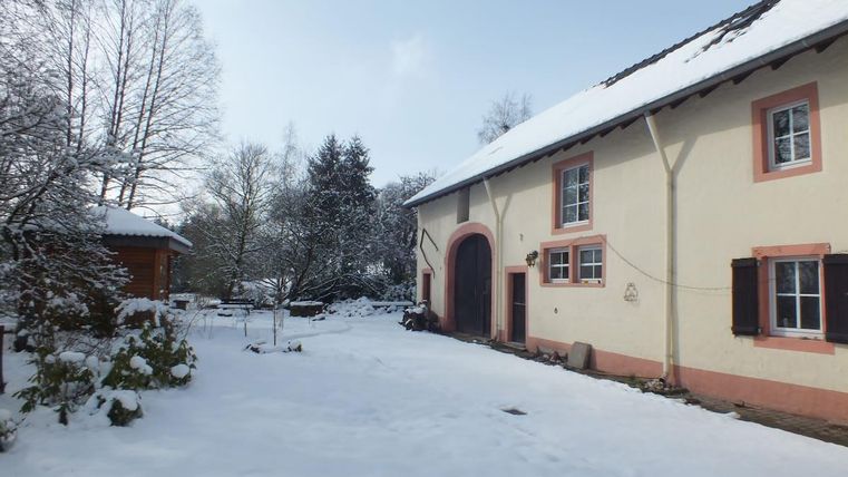 Ein charmantes Haus im Schnee mit einem roten Eingangsbereich. Die Umgebung ist mit schneebedeckten Bäumen und einem kleinen Holzhaus geschmückt.