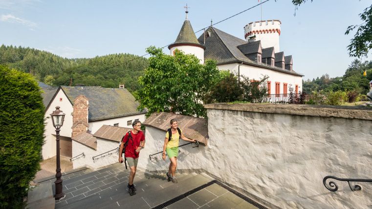 Zwei Wanderer gehen eine Treppe in Neuerburg hinauf, vorbei an einem historischen Gebäude mit Turm.