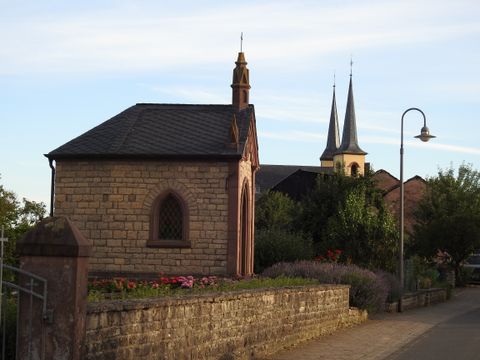 Kleine Kapelle aus Stein mit spitzem Dach, umgeben von Blumen und Bäumen, im Hintergrund Kirchtürme.