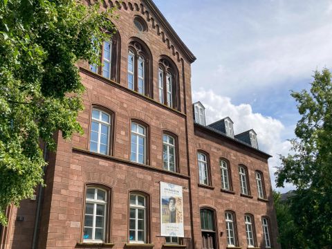 A historic building made of red stone with large windows. Surrounded by trees and a clear sky.