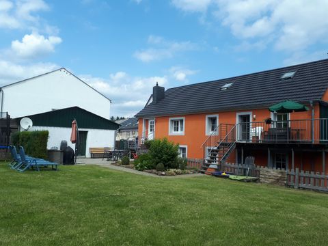 Ein schöner Garten mit einem orangefarbenen Haus und einem angrenzenden, weißen Gebäude. Es gibt Terrassenmöbel und eine Wiese mit viel Grün.