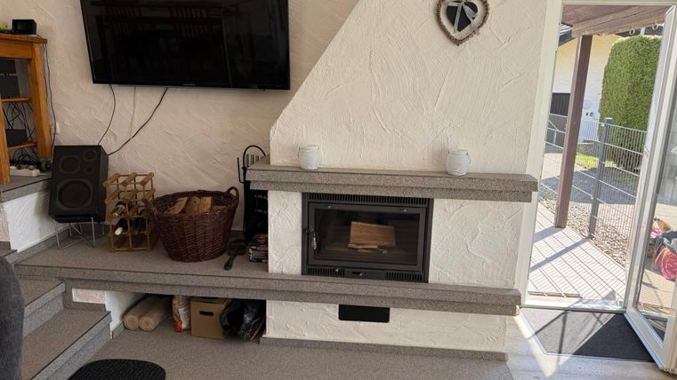 Ein gemütliches Wohnzimmer mit einem modernen Kamin und einem großen Fernseher an der Wand. Der Raum hat helle Wände und einen Zugang zur Terrasse.