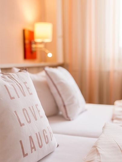 Ein gemütliches Schlafzimmer mit hellen Farben und weichen Kissen. Die Kissen tragen die Aufschrift „LIVE LOVE LAUGH“.