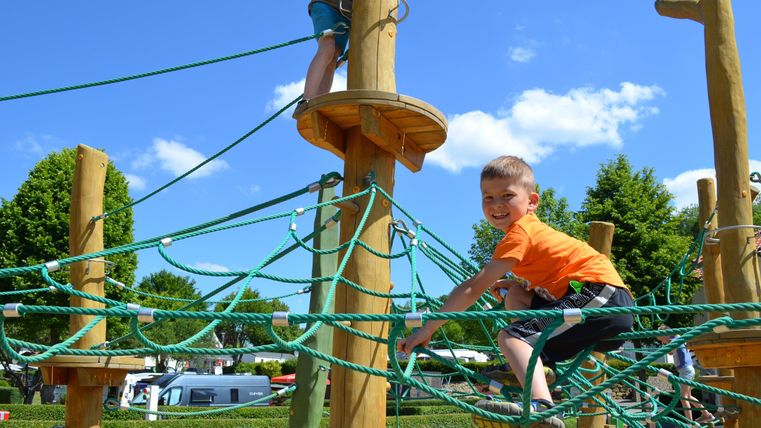 Ein Spielplatz mit einem Klettergerüst aus Holz und Seilen. Ein Junge klettert fröhlich und hat Spaß dabei.