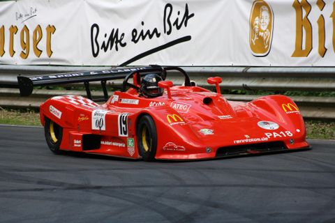 Une voiture de course rouge roule sur une piste de course. En arrière-plan, on peut voir des panneaux de sponsors.