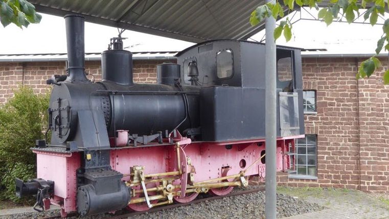 Alte Dampflokomotive unter einem Dach vor einem Backsteingebäude.